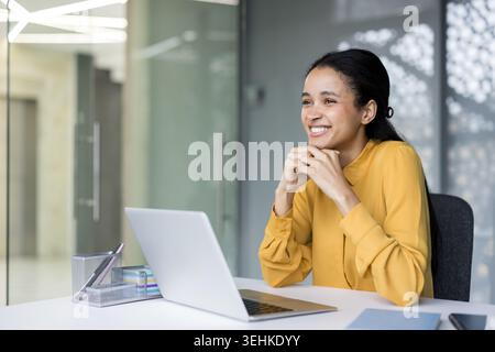 Smiling black woman professional sitting at desk with laptop in modern office, looking away and confidently dreaming about future goals and business achievement Stock Photo