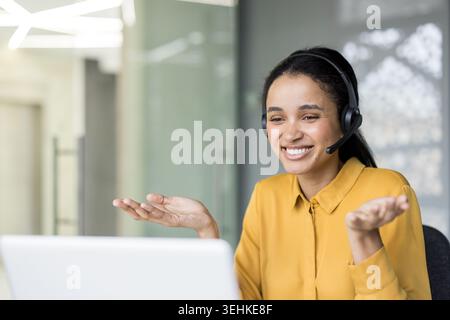 Smiling customer support agent working at a laptop in a modern office, providing online assistance and communicating with clients using a professional headset Stock Photo