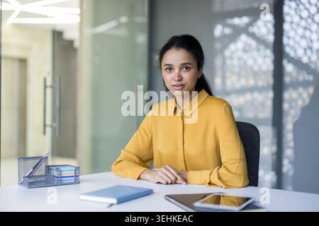Confident mixed-race businesswoman in a mustard blouse sits at a modern office desk, looking directly at camera with focused, professional expression and poised leadership presence Stock Photo