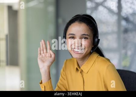 Confident female customer service agent in headset smiling and waving while offering friendly support in a modern call center, ready to assist clients via phone or online chat Stock Photo