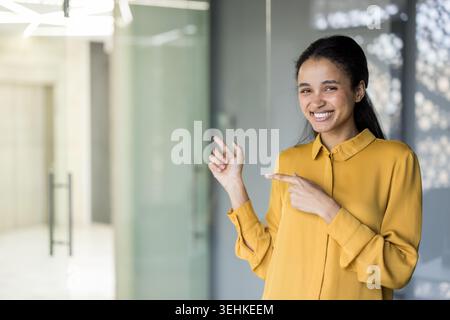 Young business professional smiling confidently in a modern office, standing and pointing sideways to introduce a product, idea, or opportunity with upbeat, approachable energy Stock Photo