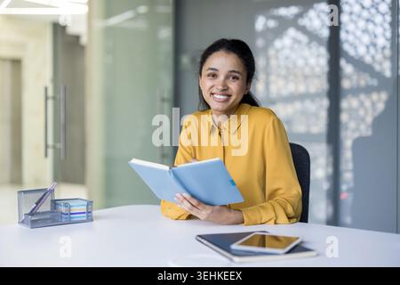 Young professional woman sitting at a modern office desk, holding a blue notebook and looking at the camera, symbolizing confidence, productivity, and a positive approach to work or study Stock Photo