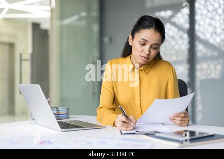 Young confident businesswoman working with concentration at her desk, holding papers, reviewing information, and writing on a clipboard in a modern office environment Stock Photo