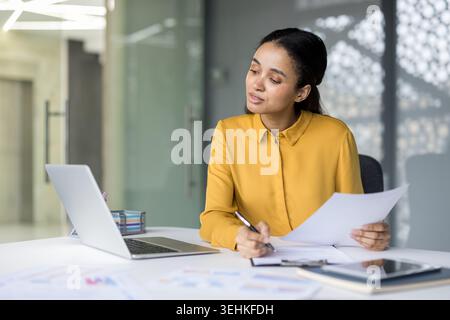 Professional woman at office desk reviewing documents and taking notes, laptop and tablet open for data analysis and planning, focused on financial reports and project strategy Stock Photo