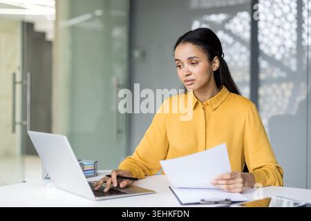 Young businesswoman with dark hair wearing a yellow shirt concentrating on her work, holding papers and using a laptop at a white desk in a modern office setup Stock Photo