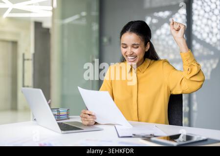 Young happy business woman in a yellow shirt celebrating professional success or positive news, reading a document at her office desk with a laptop and raising her fist in triumph Stock Photo