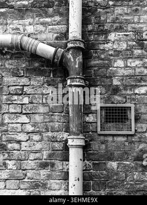 Weathered cast iron drainpipe, junction fitting and ventilation grille on a blackened Victorian brick wall, monochrome. Stock Photo