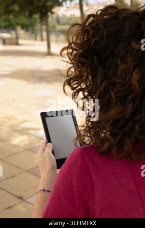 A woman leaning against a tree reads an e-book in a park with trees and benches Stock Photo