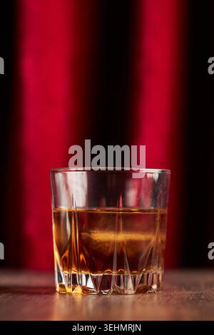 Close-up of a crystal tumbler with bourbon whisky on a wooden table, warm amber tones and reflections against a deep red curtain background, moody bar Stock Photo
