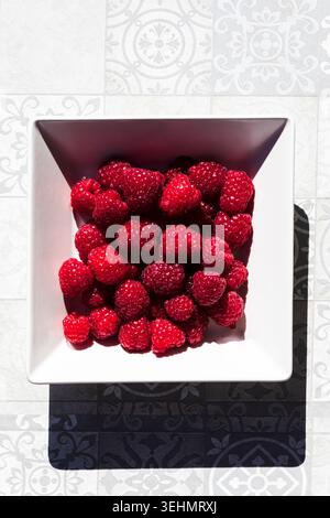 Top view of fresh raspberries piled in a square bowl, casting a bold shadow on patterned tiles; bright raspberries evoke summer, healthy eating, and c Stock Photo