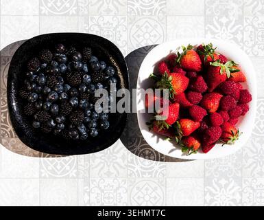 Top view of two bowls filled with fresh berries: blueberries and blackberries in a black dish and strawberries with raspberries in a white bowl on pat Stock Photo