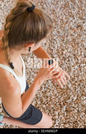 Top view of a young woman athlete checking her smart watch during an outdoor workout on gravel. Authentic lifestyle fitness moment showing resilience, Stock Photo