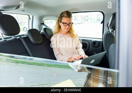 Professional business woman with glasses works on a laptop in the back seat of a car, using earbuds while traveling in the city for meetings and remot Stock Photo