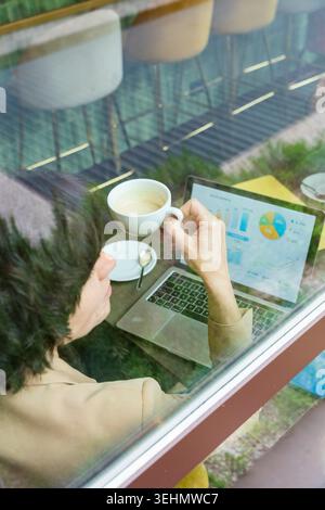 Business woman works remotely in a city cafe, holding a coffee while reviewing analytics charts on a laptop, capturing urban lifestyle, focus, and pro Stock Photo
