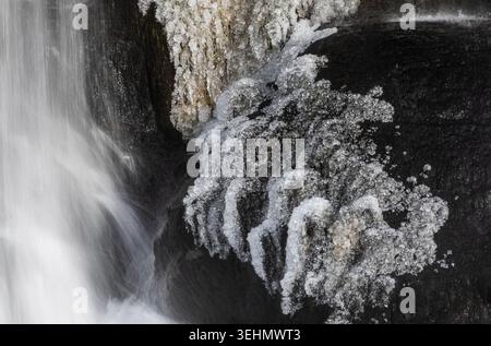 Close-up of a partially frozen waterfall, white spray and ice crystals clinging to dark volcanic rock, capturing winter texture and motion in Icelandi Stock Photo