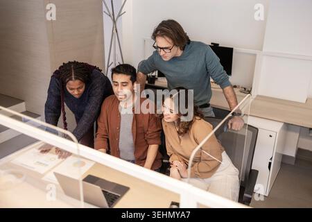 Diverse coworkers gather around a laptop in a modern coworking office, reviewing data charts and planning a project together during a focused team mee Stock Photo