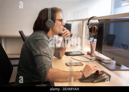 Side view of a focused coworker wearing headphones, using a mouse and monitor at a desk in a modern office, concentrating on digital work and online c Stock Photo