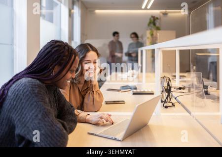 Two coworkers smiling while collaborating on a laptop at a shared desk in a modern coworking office, showing teamwork, communication, and productivity Stock Photo