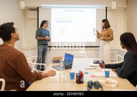 Diverse coworking team meets in a modern office as colleagues present a market analysis on a projector screen, discussing strategy, data, and next ste Stock Photo