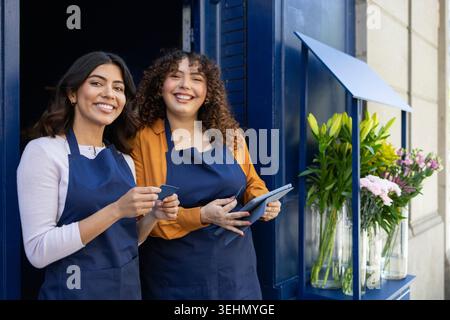 Two smiling florists stand in front of their flower shop painted in blue. The store's entrance is adorned with beautiful floral arrangements, creating Stock Photo
