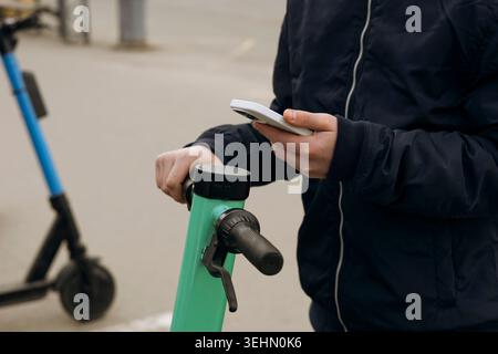 Close-up of a person in a jacket holding a smartphone while unlocking a shared electric scooter on a city street, showing modern urban mobility, renta Stock Photo
