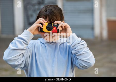 Close up portrait of a child holding a colorful disposable camera to their face outdoors, capturing a photo with flash in soft daylight and a blurred Stock Photo