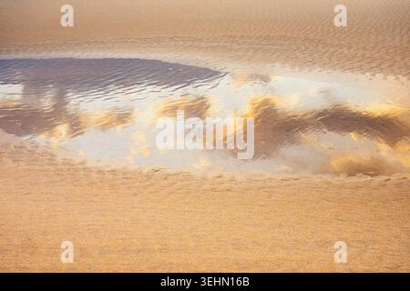 A serene beach scene featuring a shallow puddle on the sand reflecting fluffy clouds overhead. The smooth sand textures and gentle ripples create a ca Stock Photo