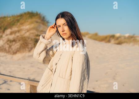 Portrait of a woman standing on a sandy beach in Aveiro, with a clear blue sky and dunes in the background. She is wearing a light coat and gently tou Stock Photo