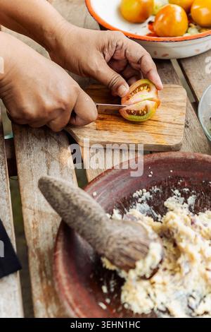 Close-up of hands slicing tomato on a wooden board beside a mortar and pestle, preparing traditional Indonesian sambal for authentic home-cooked cuisi Stock Photo