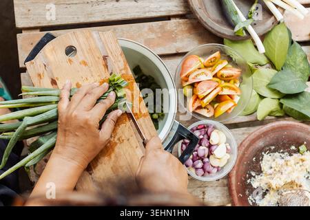 Top view of hands chopping scallions on a wooden board, surrounded by tomatoes, shallots, garlic, herbs and spices, capturing traditional Indonesian c Stock Photo