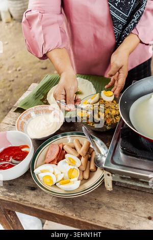 Close-up of hands serving traditional Indonesian street food with boiled eggs, sausage, sambal and dipping sauce on a rustic table, outdoor cooking in Stock Photo