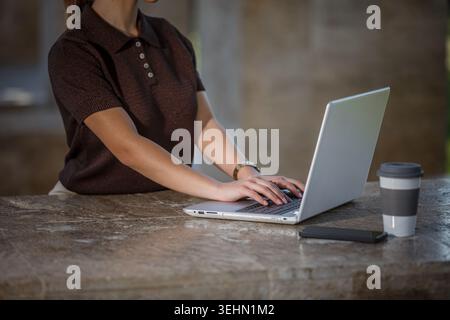 A woman works on her laptop at a stone table in a public park. She types with focused attention, surrounded by trees, a smartphone, and a cup of coffe Stock Photo