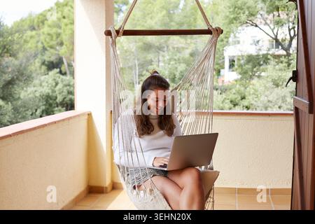 Smiling freelance woman works remotely on a laptop while sitting in a hanging chair on a sunny balcony, joining an online video call and enjoying a re Stock Photo
