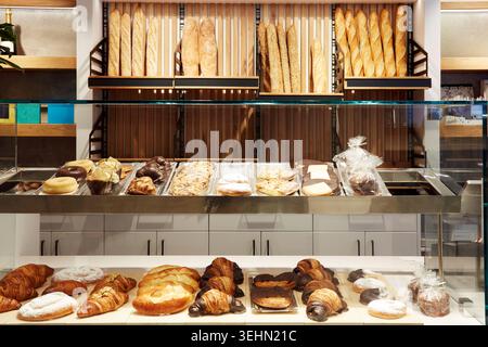 Bright modern bakery shop display with crusty baguette on wall shelves and assorted pastry, croissant, donut, and cake in glass case, inviting custome Stock Photo