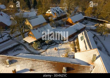 Aerial drone view of a snowy rural village in Germany at golden hour, showing farm buildings, timber houses, winter trees, and rooftops covered in fre Stock Photo