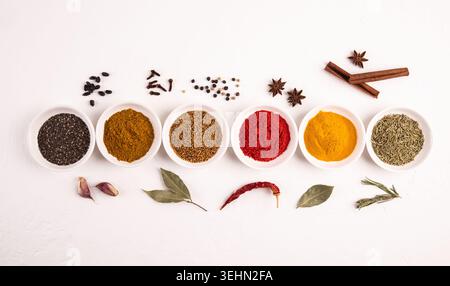 Top view of assorted spices and herbs in small bowls with pepper, turmeric, paprika, cumin, cloves, cinnamon, bay leaf, garlic, and chili on white. Stock Photo