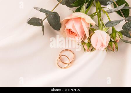 Soft flat lay of a pair of gold wedding rings beside blush pink roses and eucalyptus on a light background, symbolizing love, bride and romantic cerem Stock Photo