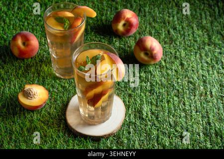 Two tall glasses filled with homemade peach ice tea, ice and mint, surrounded by fresh whole and halved peaches on a sunny lawn Stock Photo