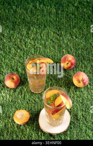 High angle view of two glasses of cold peach tea with fresh mint, scattered ripe peaches around on a green grass lawn Stock Photo