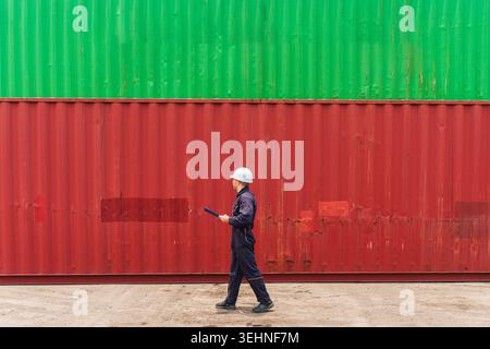 Cargo inspector caucasian man in hard hat and coveralls walking and inspecting stacked red and green shipping containers at freight port terminal Stock Photo