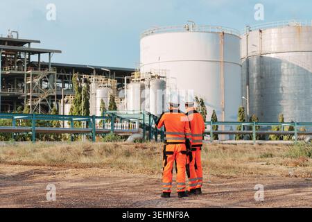 Rear view of two caucasian petrochemical engineer wearing orange high visibility safety suit with hard hat stands surveying and inspecting large metal Stock Photo