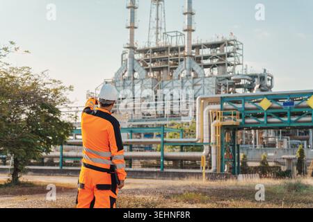 Back view of petroleum engineer caucasian man wearing orange uniform and hardhat using walkie talkie while inspecting at oil refinery plant Stock Photo