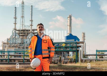 Portrait of petroleum engineer caucasian man confidently wearing safety uniform and hardhat while standing at front of industrial oil refinery network Stock Photo