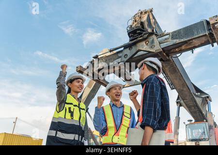 Three construction foreman workers wearing hard hat and safety vest celebrate with raised fist pump at industrial site with large crane machinery and Stock Photo