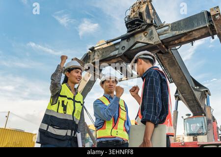 Three construction foreman workers wearing hard hat and safety vest celebrate with raised fist pump at industrial site with large crane machinery and Stock Photo
