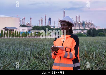 Professional black female petroleum engineer in orange high visibility jacket and white hard hat standing and using tablet at front of industrial oil Stock Photo