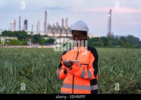 Professional black female petroleum engineer in orange high visibility jacket and white hard hat standing and using tablet at front of industrial oil Stock Photo