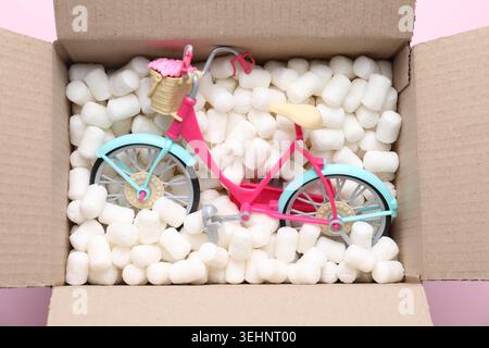Cardboard box with styrofoam pieces and toy bicycle on pink background, closeup Stock Photo