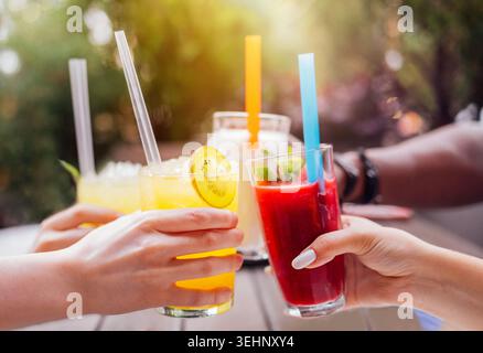 Diverse hands hold colorful cocktails with straws in a sunny outdoor setting, featuring a wooden table and blurred greenery in the background. Joyful Stock Photo
