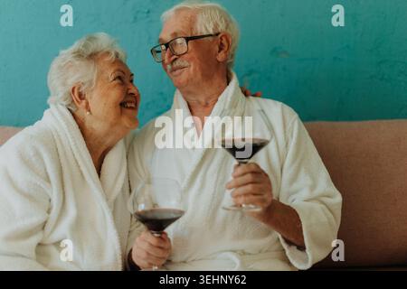 Happy senior couple sitting together in bathrobe on sofa with glass of wine. Stock Photo